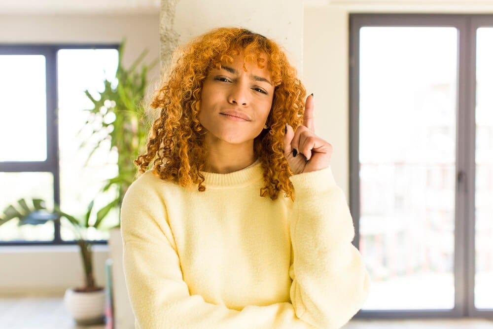 A young, attractive Latina woman with red hair smiles in a modern home.