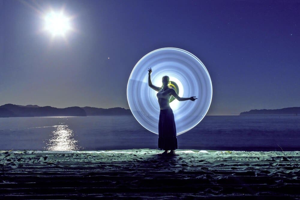 Young woman poses in front of a colorful light painting on a beach at night.