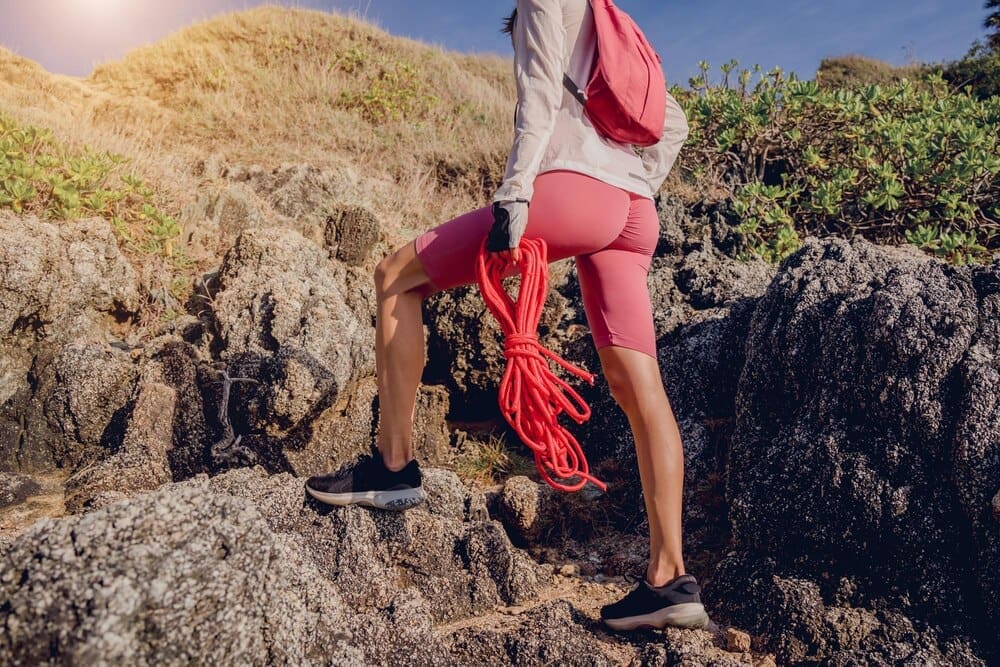 A young woman climbs a rope up a mountain, with the ocean visible in the background.