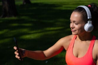 A woman in a pink top and headphones smiles while holding a phone up for a selfie