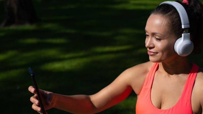 A woman in a pink top and headphones smiles while holding a phone up for a selfie