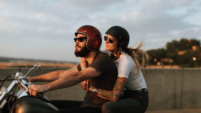 A bearded man and a woman ride a motorcycle together in the late afternoon sun
