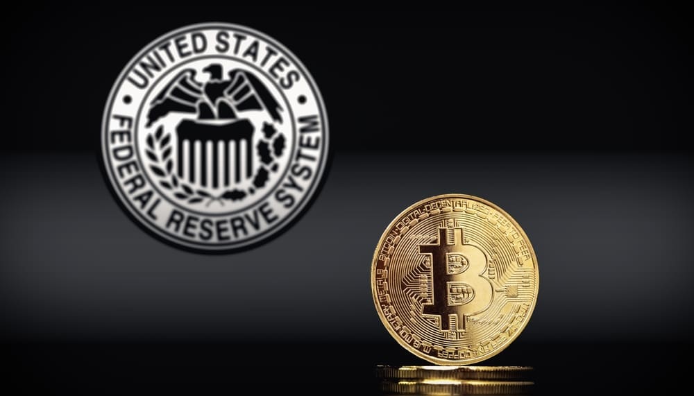 A gold Bitcoin coin stands in the foreground with the blurred seal of the U.S. Federal Reserve System in the background