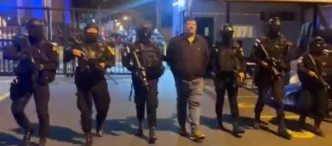 A man in a polo shirt and jeans is being escorted by a group of heavily armed and masked police officers at night