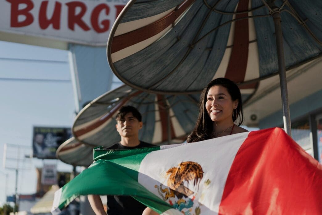 A smiling woman holds a large Mexican flag with a man standing behind her, celebrating a holiday in the United States