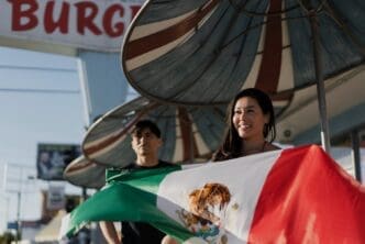 A smiling woman holds a large Mexican flag with a man standing behind her, celebrating a holiday in the United States