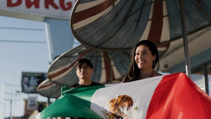 A smiling woman holds a large Mexican flag with a man standing behind her, celebrating a holiday in the United States