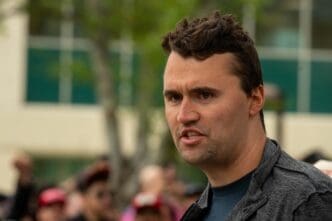 A man with short, brown hair wearing a dark jacket speaks with his mouth slightly open