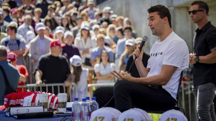 Charlie Kirk is seated and speaking into a microphone with a large crowd in the background at an outdoor event