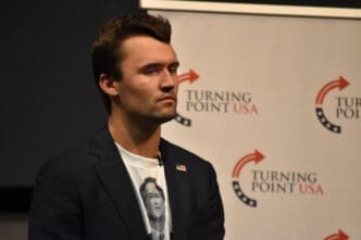 Charlie Kirk stands with a serious expression in front of a Turning Point USA banner