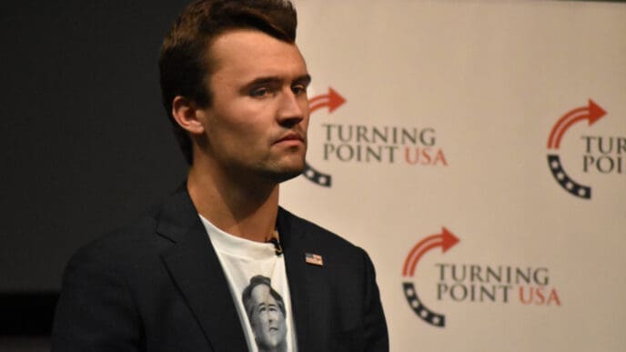 Charlie Kirk stands with a serious expression in front of a Turning Point USA banner