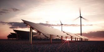 Rows of solar panels and two wind turbines at sunset, with the sun's rays peaking through the panels