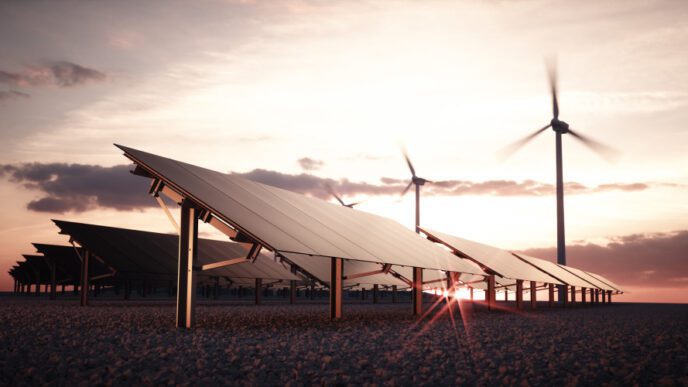 Rows of solar panels and two wind turbines at sunset, with the sun's rays peaking through the panels