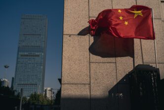 The red Chinese flag flutters in the wind next to a modern stone building and a skyscraper