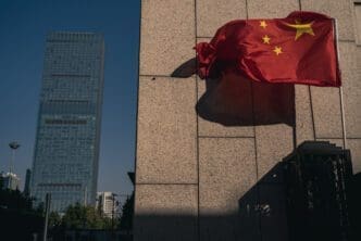 The red Chinese flag flutters in the wind next to a modern stone building and a skyscraper