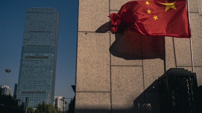 The red Chinese flag flutters in the wind next to a modern stone building and a skyscraper