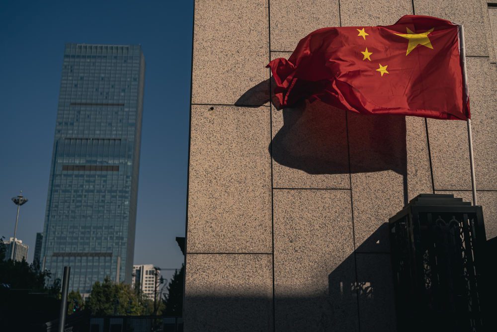 The red Chinese flag flutters in the wind next to a modern stone building and a skyscraper
