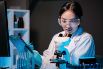 A Chinese scientist in a lab coat and safety goggles, looking at a clipboard near a microscope