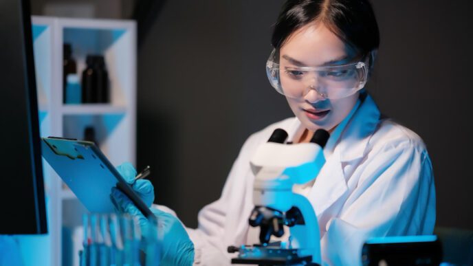 A Chinese scientist in a lab coat and safety goggles, looking at a clipboard near a microscope
