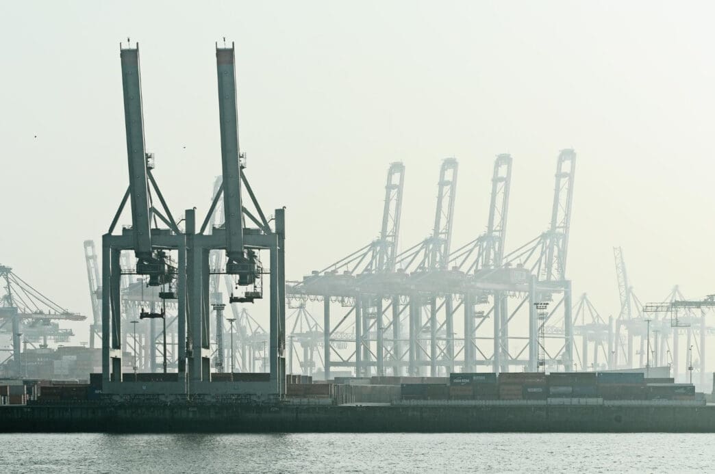 A line of immense cranes and stacked shipping containers are silhouetted in the haze of a large cargo port