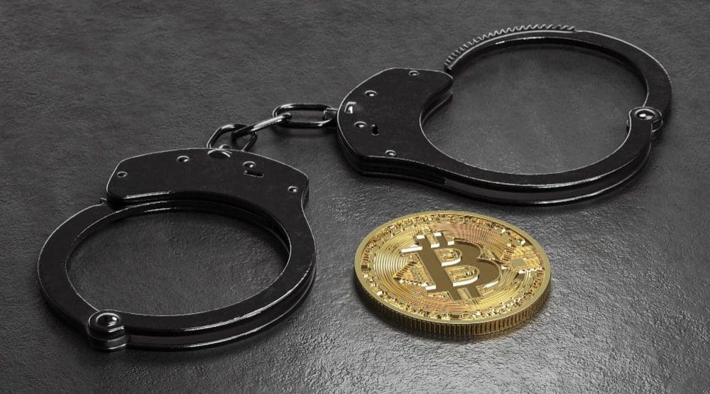 A pair of black handcuffs and a shiny golden Bitcoin coin on a dark, textured surface
