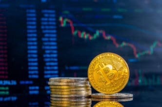A physical Bitcoin coin and a stack of coins are shown in front of a blurred digital trading chart