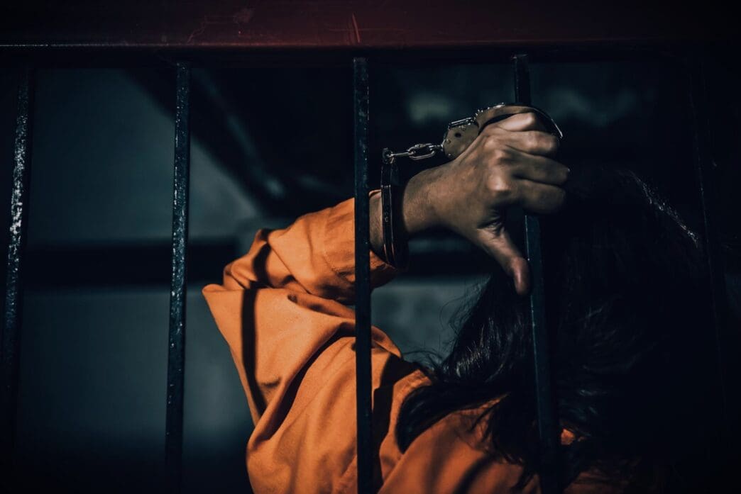 A person in an orange jumpsuit with their cuffed hands clinging to the iron bars of a prison cell, conveying a feeling of despair