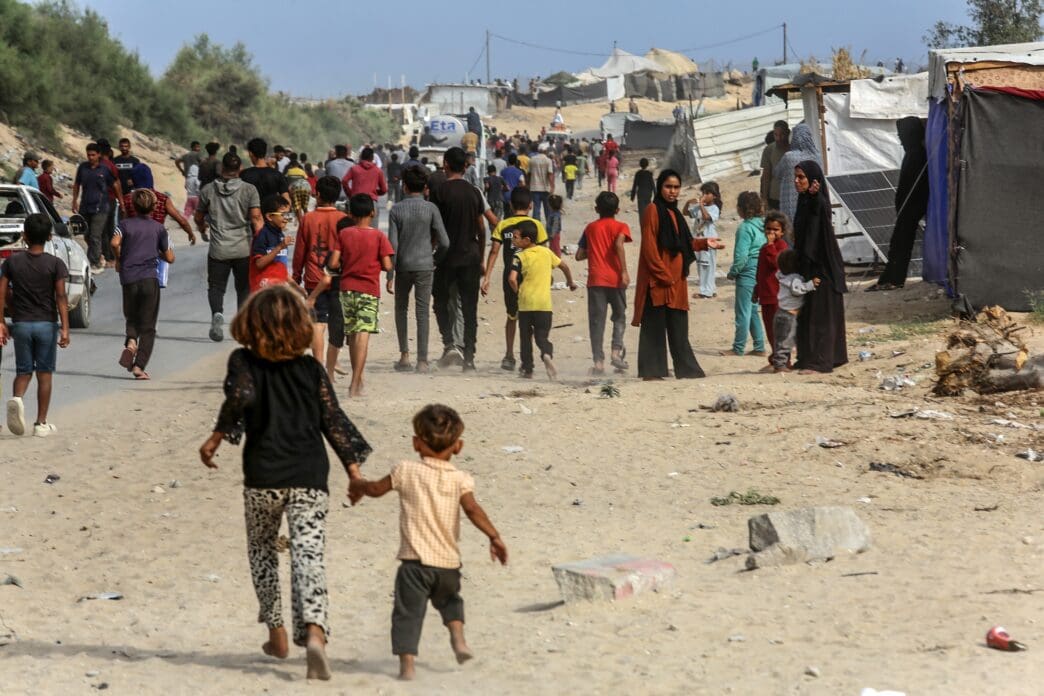 Displaced people walking through a sandy area with tents and damaged buildings in Gaza