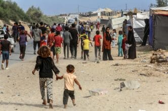 Displaced people walking through a sandy area with tents and damaged buildings in Gaza