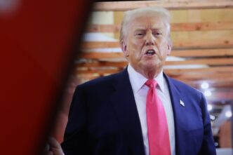 Donald Trump speaking and gesturing with a serious expression, wearing a navy suit and a pink tie
