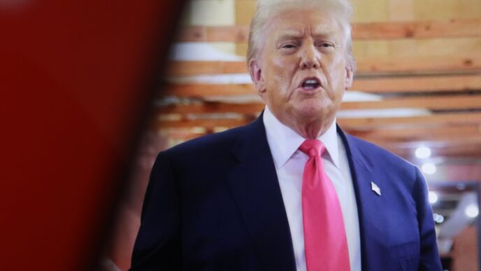 Donald Trump speaking and gesturing with a serious expression, wearing a navy suit and a pink tie