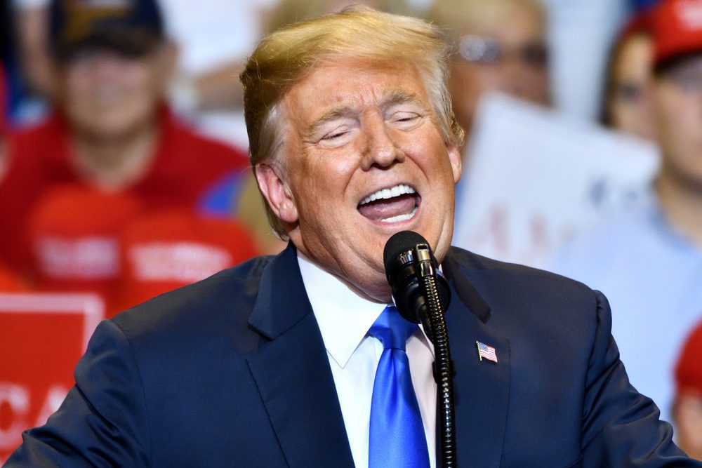 Donald Trump speaking at a rally, with his eyes closed and mouth open, in front of a crowd