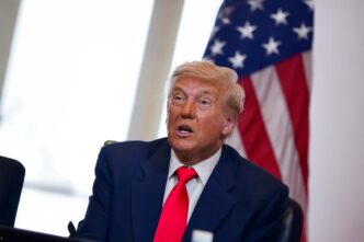 Donald Trump seated at a table, looking up and speaking with a US flag in the background