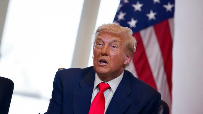 Donald Trump seated at a table, looking up and speaking with a US flag in the background