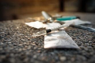 A syringe and a small bag of a white substance are shown on a rough surface, representing illegal drug use