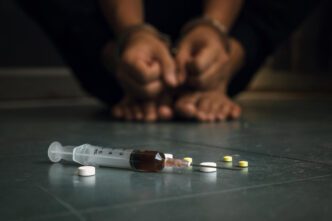 A syringe and pills on a floor, with a person in handcuffs blurred in the background