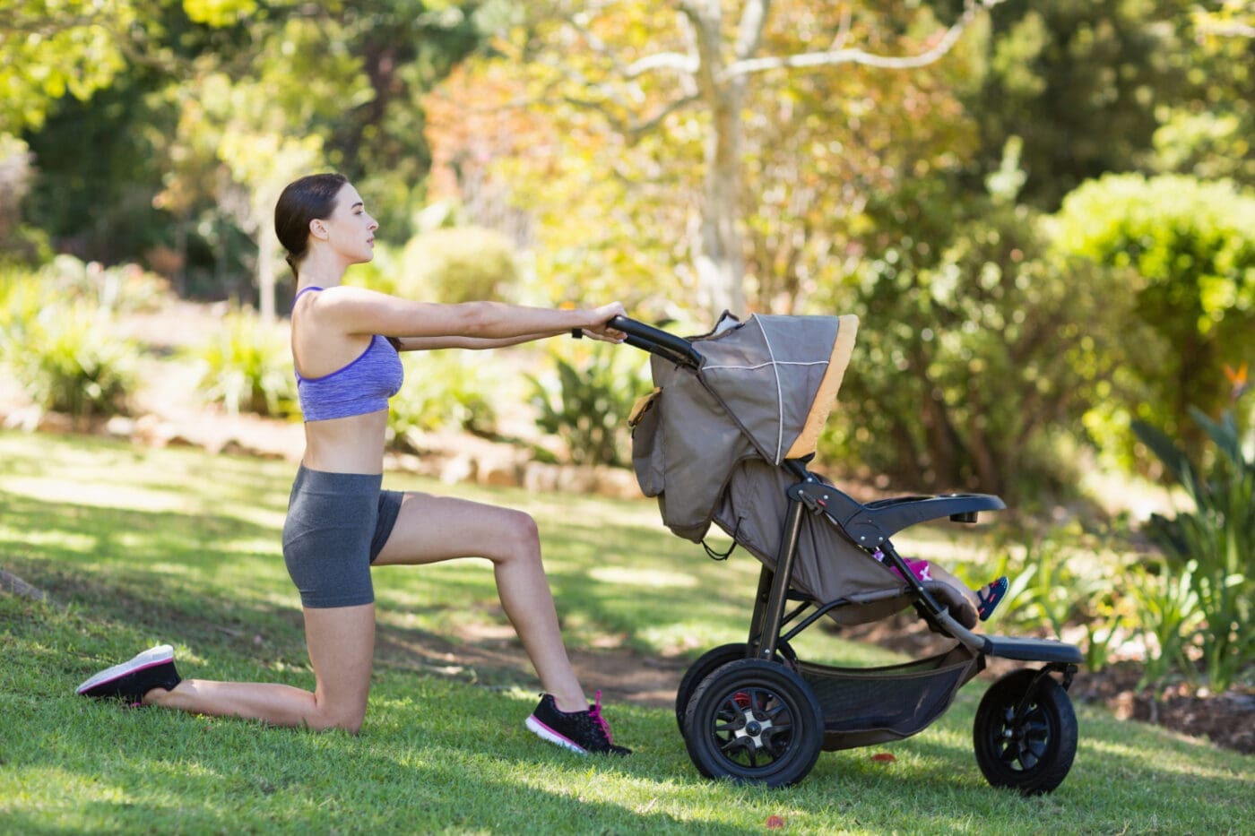 A young woman performs a lunge while using a baby stroller for balance in a sunny park