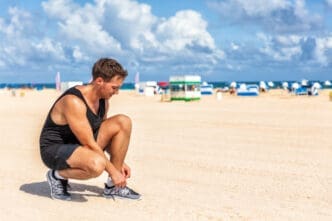 A man in a black tank top and shorts squats on a beach to tie his running shoe