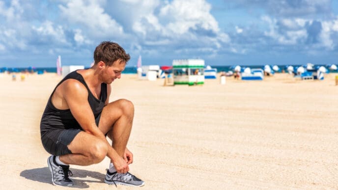 A man in a black tank top and shorts squats on a beach to tie his running shoe