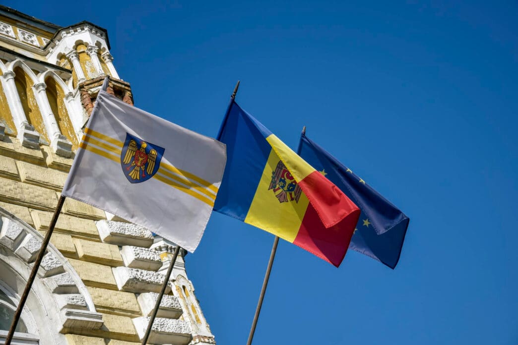 Three flags, including Moldova and the EU, fly against a blue sky near a building