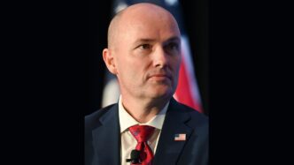 Governor Spencer Cox of Utah, a bald man in a suit, looks thoughtfully away from the camera
