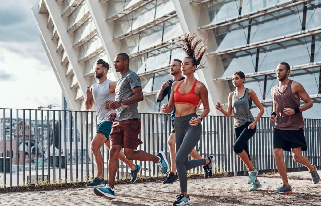 A diverse group of six friends jogging together on a concrete path next to a modern building