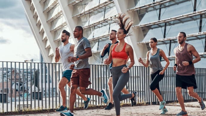A diverse group of six friends jogging together on a concrete path next to a modern building
