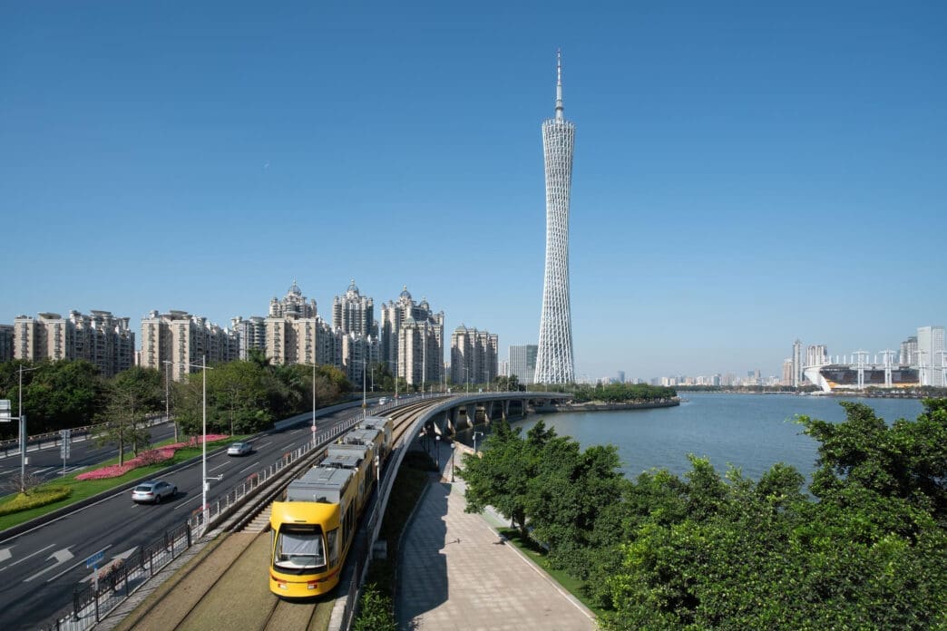 A yellow tram passes by a tower and cityscape in Guangzhou under a clear blue sky.