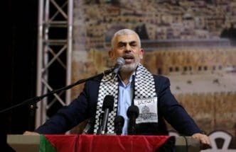 Hamas leader Yahya Sinwar speaking at a conference with a crowd and city in the background