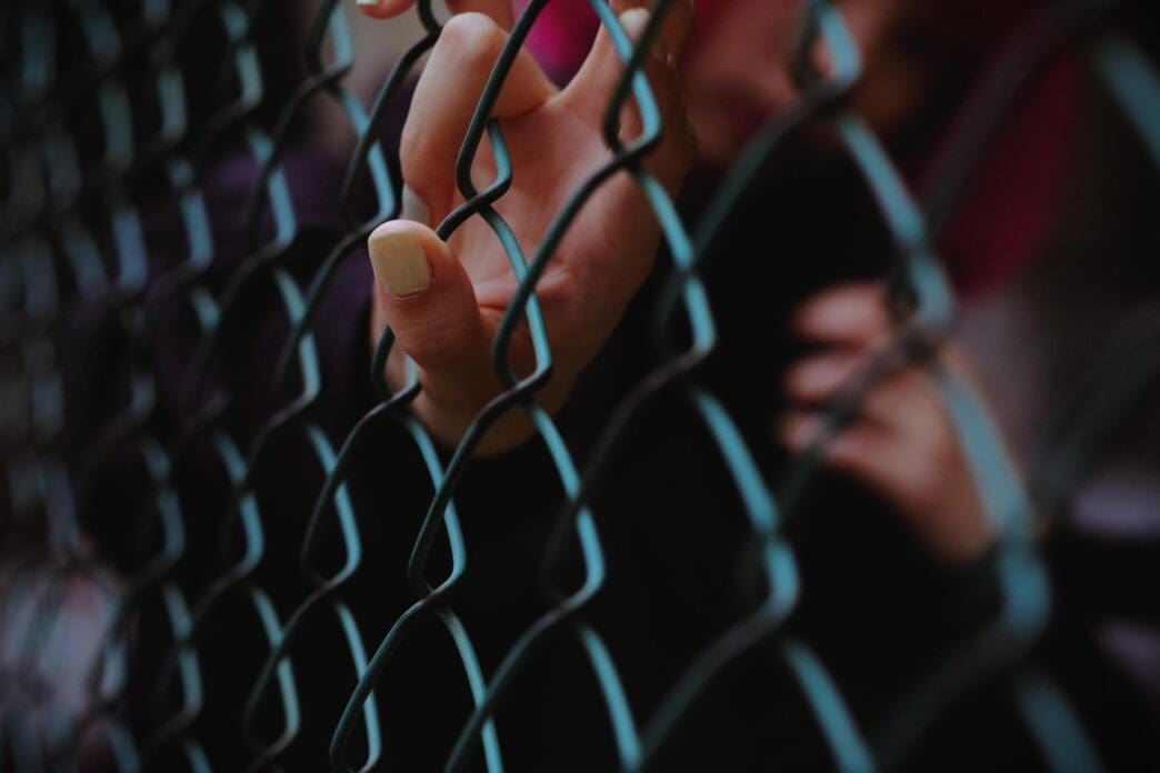 Hand with pale yellow nail polish touching a chain-link fence