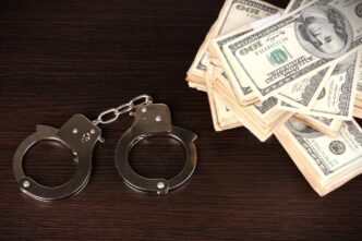A pair of handcuffs lying next to a large stack of US hundred-dollar bills on a dark wooden surface.