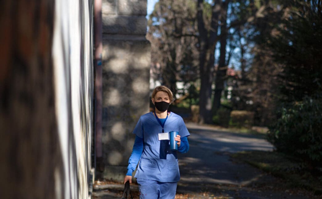 A female healthcare worker in scrubs and a mask walks outdoors with a cup