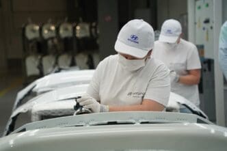 Two factory workers in masks and hats work on a white car part at a Hyundai plant