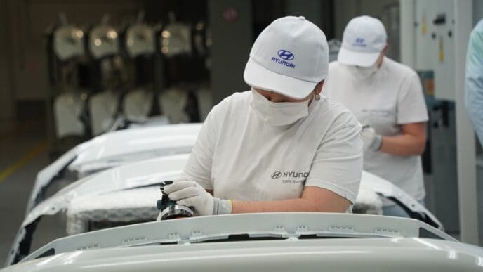 Two factory workers in masks and hats work on a white car part at a Hyundai plant